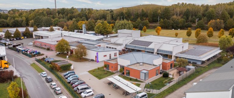 Werkstatt für angepasste Arbeit Die Werkstatt. Luftaufnahme der Werkstatt und umliegenden Parkplätzen. Viele bunte Bäume.