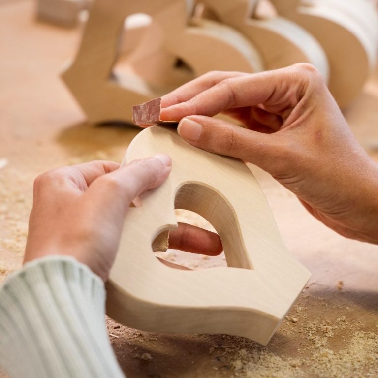 Fertigung Deko Herz Holzherz. Hände schleifen ein herzförmiges Holzstück, umgeben von weiteren Holzherzen.